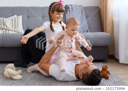 Portrait of brunette father playing with his daughters while lying on floor near sofa, wearing white t shirt, children sitting on dad and laughing, happy childhood. 102173994