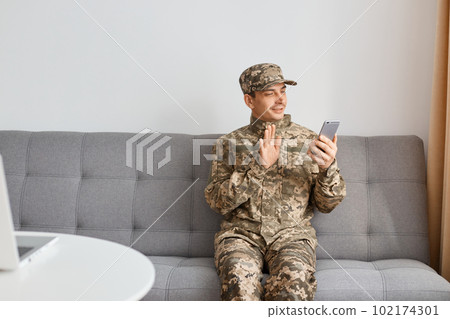 Indoor shot of young adult military man wearing camouflage uniform sitting on sofa, soldier sitting with mobile phone in hands, waving palm while having video call. Indoor shot of young adult military man wearing camouflage uniform sitting on sofa, soldier sitting with mobile phone in hands, waving palm while having video call. 102174301