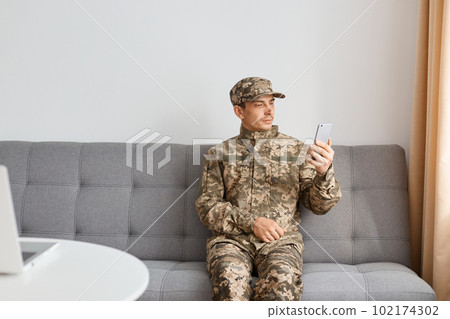 Horizontal shot of Caucasian military man wearing camouflage uniform sitting on sofa, soldier holding cell phone, looking at display, watching video or having online conversation. Horizontal shot of Caucasian military man wearing camouflage uniform sitting on sofa, soldier holding cell phone, looking at display, watching video or having online conversation. 102174302