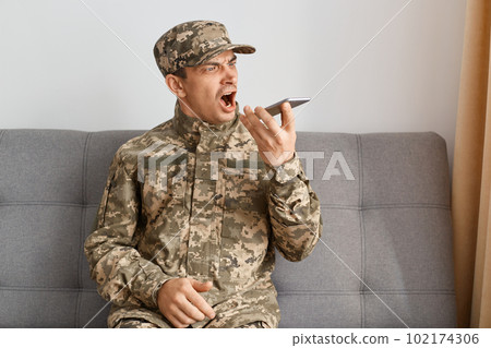 Indoor shot of angry military man wearing camouflage uniform sitting on sofa, soldier recording voice message with aggressive emotions, screaming with anger and hate. 102174306