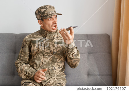 Image of aggressive military man wearing camouflage uniform sitting on sofa, soldier using voice assistant or recording voice message, yelling, being very angry. Image of aggressive military man wearing camouflage uniform sitting on sofa, soldier using voice assistant or recording voice message, yelling, being very angry. 102174307