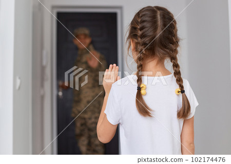 Indoor shot of military dad goes to war, little girl with dark hair and braids posing backwards and waving hand, saying good bye to father posing at the door. Indoor shot of military dad goes to war, little girl with dark hair and braids posing backwards and waving hand, saying good bye to father posing at the door. 102174476