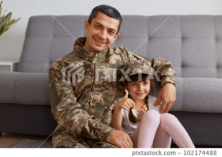 Portrait of smiling satisfied young adult soldier man wearing camouflage uniform returning home after army, spending time with daughter, looking at camera with happy expression, kid showing thumb up. 102174488