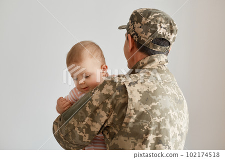 Back view of military father came from the war or the exercises and holding his infant baby, missing his cute toddler daughter, wearing camouflage uniform and cap. Back view of military father came from the war or the exercises and holding his infant baby, missing his cute toddler daughter, wearing camouflage uniform and cap. 102174518