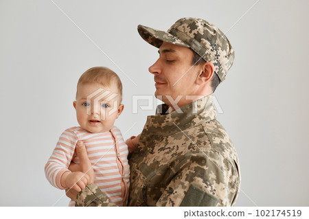 Side view of young adult military father came from the war or the exercises and holding hid cute baby girl wearing striped sleeper, armyman wearing camouflage uniform and cap. Side view of young adult military father came from the war or the exercises and holding hid cute baby girl wearing striped sleeper, armyman wearing camouflage uniform and cap. 102174519