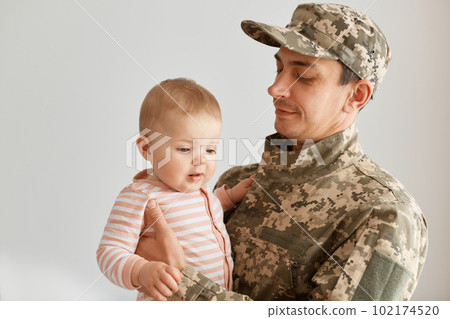 Indoor shot of military father came from the war or the exercises and posing with his little toddler daughter in striped sleeper, posing at home after returning home. Indoor shot of military father came from the war or the exercises and posing with his little toddler daughter in striped sleeper, posing at home after returning home. 102174520