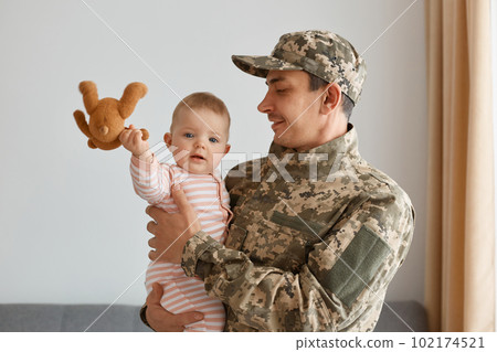 Indoor shot of smiling military man wearing camouflage uniform and cap, standing with his little infant daughter in hands, baby holding soft toy, being glad to play with father. Indoor shot of smiling military man wearing camouflage uniform and cap, standing with his little infant daughter in hands, baby holding soft toy, being glad to play with father. 102174521