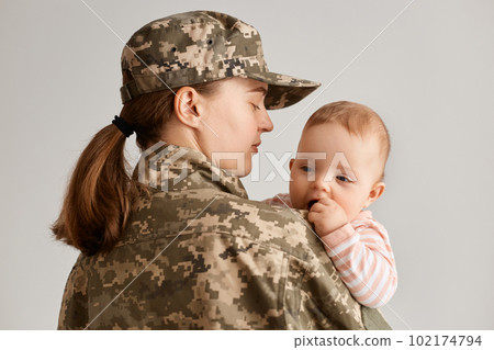 Indoor shot of young adult woman soldier posing backwards to camera with infant baby in hands, military mother returning home from army or war, being glad to see her little daughter. 102174794