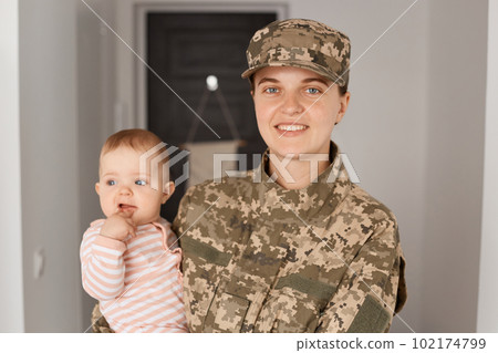 Portrait of happy smiling young adult woman soldier wearing camouflage uniform, returning home and being glad to meet her family, holding toddler girl in arms. 102174799