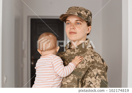 Happy positive optimistic proud young adult woman soldier wearing camouflage uniform, returning home and taking her little daughter, looking away with glad expression. 102174819