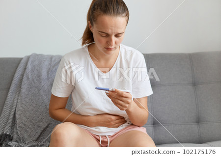 Sad unhappy woman wearing while casual style t shirt waiting for pregnancy test result thinking about future, touching her belly while sitting on sofa in living room alone. Sad unhappy woman wearing while casual style t shirt waiting for pregnancy test result thinking about future, touching her belly while sitting on sofa in living room alone. 102175176