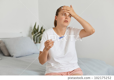 Sad unhappy young adult pregnant woman with pregnancy test posing in her bedroom, wearing white t shirt, looking away with sorrow expression, keeping hand on forehead. Sad unhappy young adult pregnant woman with pregnancy test posing in her bedroom, wearing white t shirt, looking away with sorrow expression, keeping hand on forehead. 102175252
