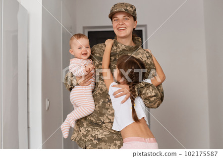 Indoor shot of positive young adult woman soldier wearing camouflage uniform, children hugging her heroic mother while returning home, female looking at camera with happy expression. 102175587