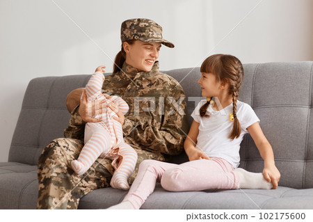 Portrait of attractive positive soldier woman wearing camouflage uniform sitting on sofa with her kids and talking, missing her children so much while being in army or war. Portrait of attractive positive soldier woman wearing camouflage uniform sitting on sofa with her kids and talking, missing her children so much while being in army or war. 102175600