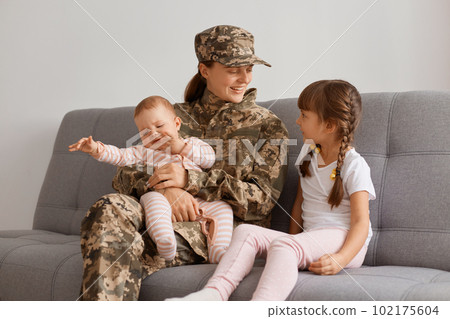 Portrait of smiling soldier woman wearing camouflage uniform sitting on sofa with kids, being extremely happy to see her children after Military service, motherhood. 102175604