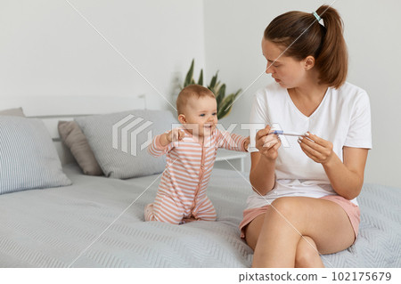 Indoor shot of attractive female with pigtail holding pregnancy test in hands, sitting on bed in bedroom with her infant daughter, looking at her baby, waiting result. Indoor shot of attractive female with pigtail holding pregnancy test in hands, sitting on bed in bedroom with her infant daughter, looking at her baby, waiting result. 102175679