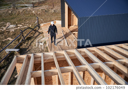 Male engineer building wooden frame house. Man developer on construction site, inspecting quality of work on sunny day, holding hammer in hands. Male engineer building wooden frame house. Man developer on construction site, inspecting quality of work on sunny day, holding hammer in hands. 102176049