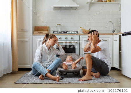 Portrait of mother and father sitting on floor in kitchen with little son or daughter in rocking chair on kitchen floor, tired sleepless parents keeping hands on head, need rest. 102176191