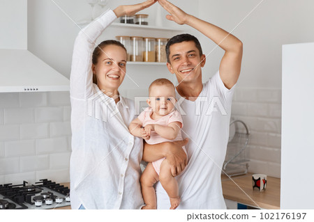 Indoor shot of happy smiling couple standing with baby daughter and making roof with hands above their heads, feels in safety together at home, expressing positive emotions. 102176197