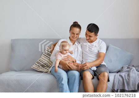 Indoor shot of happy mother, father with baby daughter at home, sitting on sofa, spending time together, expressing love, gentle and happiness, family with cute child. Indoor shot of happy mother, father with baby daughter at home, sitting on sofa, spending time together, expressing love, gentle and happiness, family with cute child. 102176218