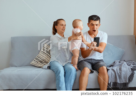 Indoor shot of young parents with their kid sitting on cough in light room, man holding cell phone, being shocked to win, betting, man screaming with amazement, woman giving baby to father. 102176235