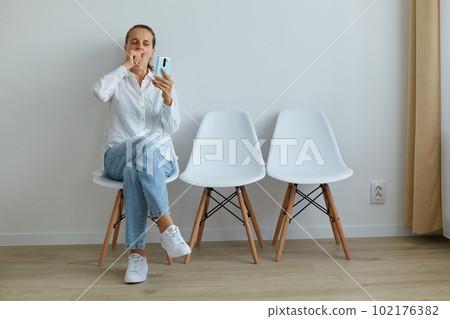 Indoor shot of tired bored female sitting in queue on chair against light wall, wearing casual style clothing, covering mouth while yawning, using phone while long time waiting. 102176382