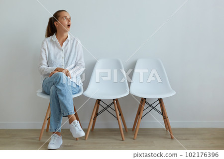 Astonished female wearing casual clothing sitting on chair and looking away with widely open mouth, sees something shocked, being impressed by surprised things. Astonished female wearing casual clothing sitting on chair and looking away with widely open mouth, sees something shocked, being impressed by surprised things. 102176396