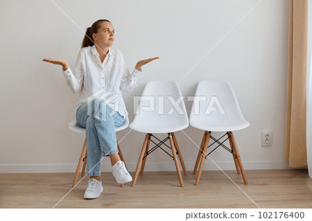 Horizontal shot of helpless confused woman wearing white shirt and jeans sitting on chair in queue, spreading hands, shrugging shoulders, expressing puzzlement. Horizontal shot of helpless confused woman wearing white shirt and jeans sitting on chair in queue, spreading hands, shrugging shoulders, expressing puzzlement. 102176400