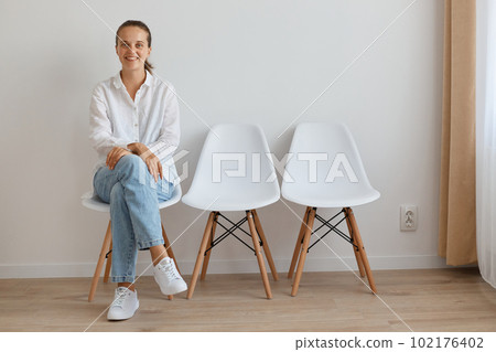 Portrait of happy smiling woman wearing white shirt and jeans sitting on chair in queue, holding her hands on her legs, looking at camera with optimistic expression. 102176402