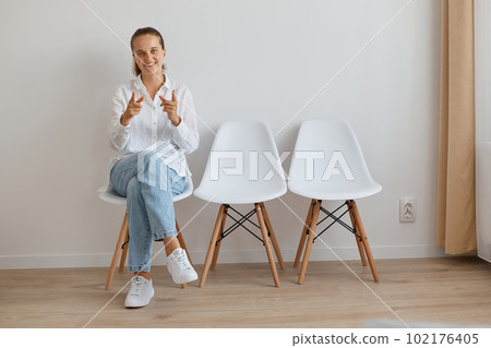 Full length portrait of satisfied young adult woman wearing white shirt and jeans sitting on chair in queue, looking and pointing at camera, being happy and delighted. 102176405