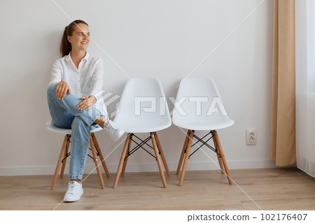 elf confident beautiful woman with ponytail wearing white shirt and jeans sitting on chair in queue, looks calm, looking away with smile, expressing positive emotions. 102176407