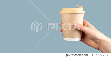Female hand holding a paper cup of coffee over pastel blue background, banner, copy space. Disposable takeaway takeout mug of latte with lid cover 102177287
