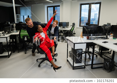 Caucasian red-haired woman, bearded caucasian man rolled African American young woman on office chair. Colleagues have fun at work. Caucasian red-haired woman, bearded caucasian man rolled African American young woman on office chair. Colleagues have fun at work. 102178201