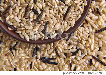 A mixture of brown and black unpolished wild rice in a clay ceramic bowl close-up against the background of scattered rice grains 102178788