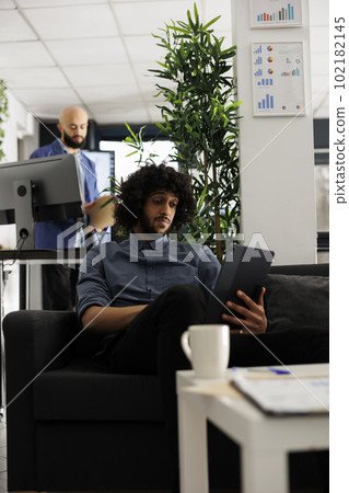 Unicron business professional using digital table during work break in start up office. Entrepreneur watching video on portable gadget while sitting on couch in modern coworking space 102182145