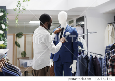 Clothing store stylist creating formal wear outfit on mannequin in retail outlet. African american man dressing dummy model in suit and fixing tie while working in shopping mall 102182179