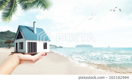 Moving to island. Female hand holding 3D model of small comfortable house against beach, palms and ocean background 102182994