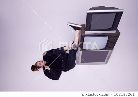Top view. Beautiful young girl in jeans clothes lying on floor near retro tv sets over grey studio background. News, information Top view. Beautiful young girl in jeans clothes lying on floor near retro tv sets over grey studio background. News, information 102183261