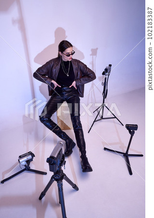 Portrait of young beautiful girl posing in stylish leather jacket, pants and trendy sunglasses over retro cameras Portrait of young beautiful girl posing in stylish leather jacket, pants and trendy sunglasses over retro cameras 102183387