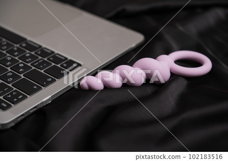 Close-up of lilac anal beads next to laptop on black silk sheet. Close-up of lilac anal beads next to laptop on black silk sheet. 102183516
