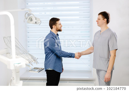 The doctor meets patient. Man shaking hands with dentist standing near dentist chair in office. 102184770