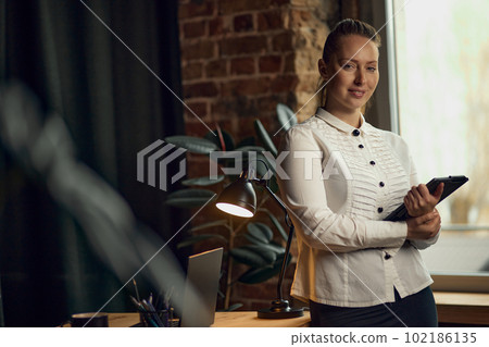 Portrait of successful, confident, independent business woman standing near window with table and looking at camera 102186135