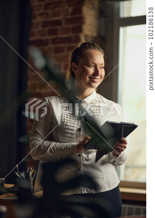 Portrait of young, smiling, beautiful business woman standing near window, looking outside, working on tablet in her office Portrait of young, smiling, beautiful business woman standing near window, looking outside, working on tablet in her office 102186138