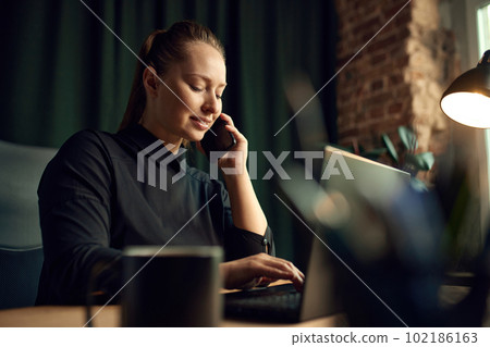 Beautiful female employee, business woman sitting at her office at table, talking on mobile phone, working on laptop 102186163