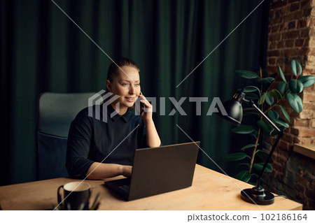 Smiling, calm, happy business woman sitting t table with laptop and talking on mobile phone. Good news, deal, partnership 102186164