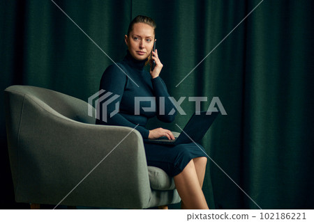 Confident, beautiful, independent business woman sitting on armchair in her office, working on laptop and talking on mobile phone 102186221