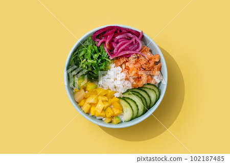 Poke bowl with rice, salmon,cucumber,mango,onion,wakame salad, poppy seeds ands sunflowers seeds isolated on yellow background.. Poke bowl with rice, salmon,cucumber,mango,onion,wakame salad, poppy seeds ands sunflowers seeds isolated on yellow background.. 102187485