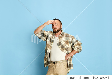 Portrait of bearded man in casual clothes holding hand to forehead, showing shocked emotions against blue studio background Portrait of bearded man in casual clothes holding hand to forehead, showing shocked emotions against blue studio background 102189051