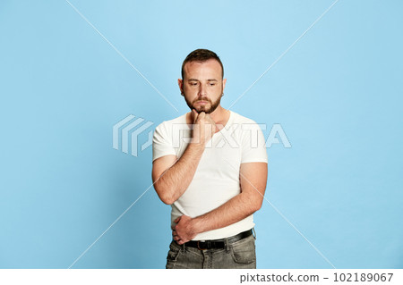 Portrait of bearded man in casual clothes posing, standing with sad, thoughtful, depressed face against blue studio background 102189067