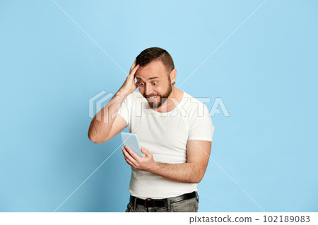 Portrait of happy, excited, bearded man in white t-shirt emotionally looking on mobile phone with shocked, positive face against blue studio background Portrait of happy, excited, bearded man in white t-shirt emotionally looking on mobile phone with shocked, positive face against blue studio background 102189083